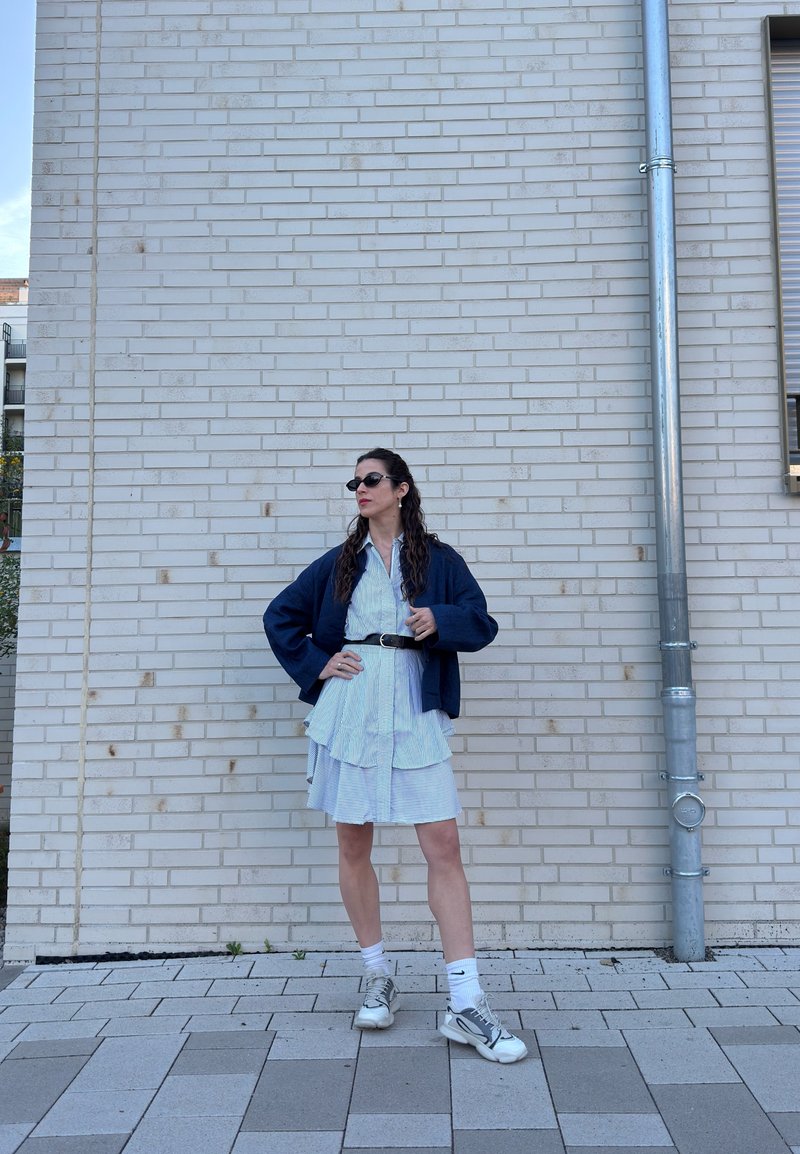 Frau mit Sonnenbrille, blauer Jacke, gestreiftem Kleid, weißen Socken und Turnschuhen steht an einer hellen Ziegelwand neben einem Metallrohr.