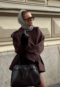 Bruine trenchcoat met oversized zonnebril, grijze sjaal, zwarte leren handschoenen, houdt een zwarte handtas met gouden accenten vast, tegen een lichte muur.
