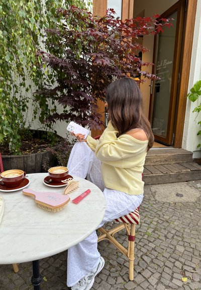 Dos tazas marrones de café con leche sobre una mesa de mármol con un cepillo de pelo rosa, un producto labial y un teléfono en la mano de una persona; entorno exterior con vegetación.