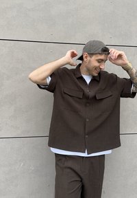 Brązowa koszula z krótkim rękawem z dwoma kieszeniami na piersi, w połączeniu z pasującymi spodniami. Biała podkoszulka i szara czapka. Gładka, teksturowana tkanina.