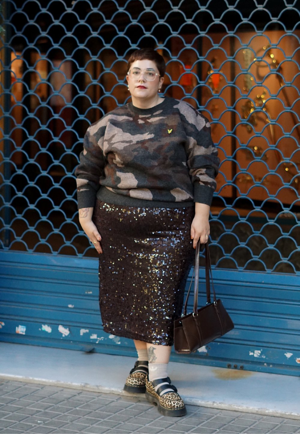Personne aux cheveux courts portant un pull camouflage, une jupe à paillettes, des chaussures à imprimé léopard, tenant un sac à main marron, debout devant une vitrine bleue fermée.