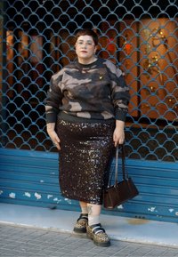 Personne aux cheveux courts portant un pull camouflage, une jupe à paillettes, des chaussures à imprimé léopard, tenant un sac à main marron, debout devant une vitrine bleue fermée.