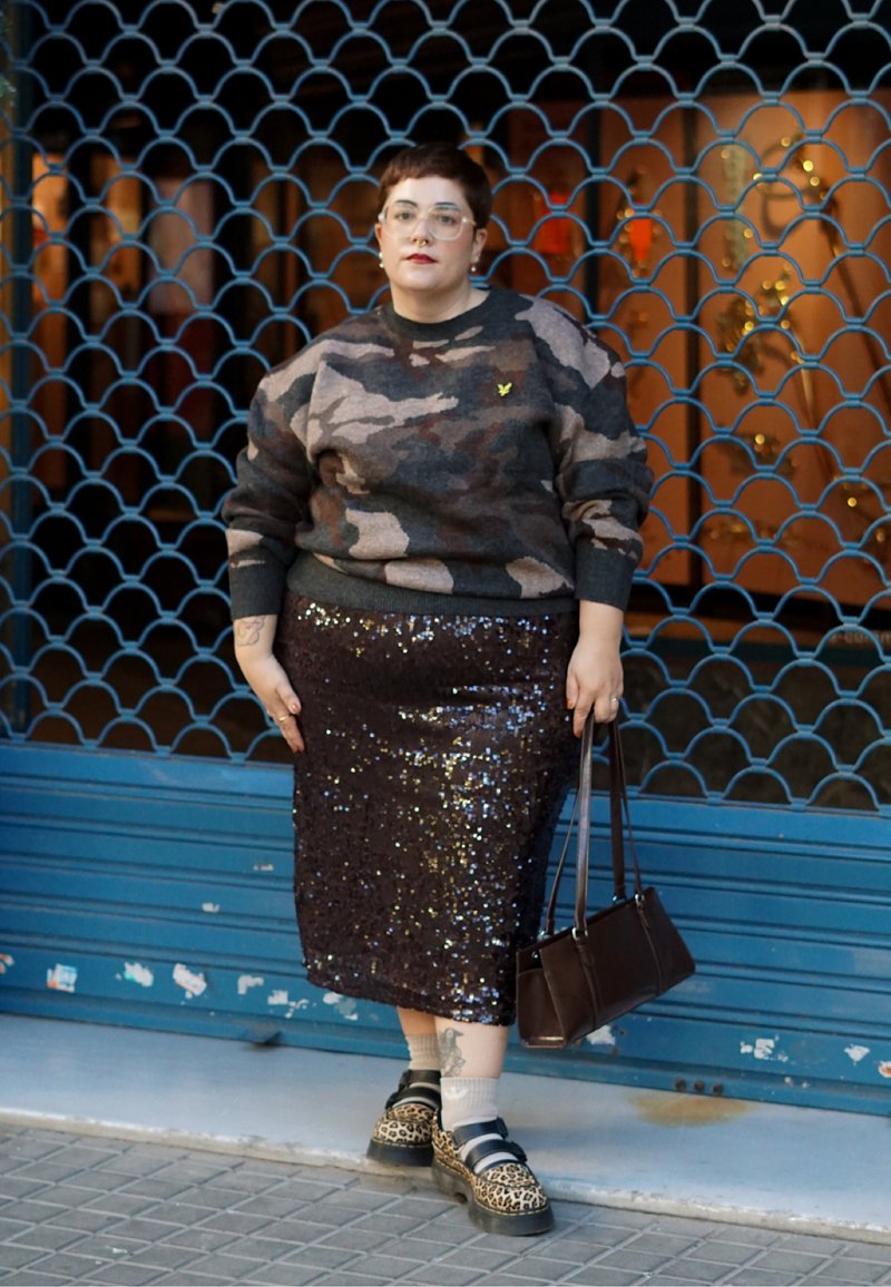 Άτομο με κοντά μαλλιά που φοράει πουλόβερ παραλλαγής, φούστα με παγιέτες, παπούτσια με λεοπάρ μοτίβο, κρατώντας καφέ τσάντα, στέκεται μπροστά σε κλειστή μπλε βιτρίνα.