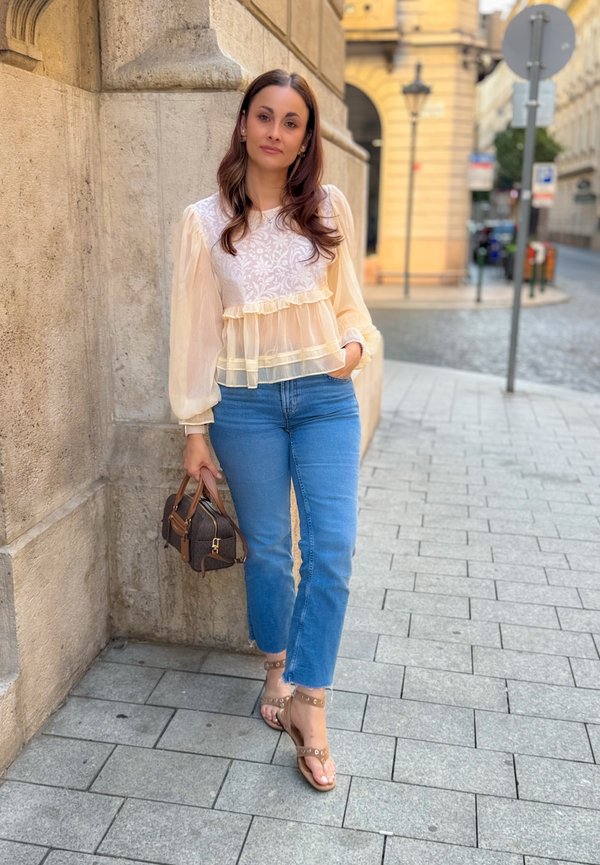 Mujer con blusa crema transparente, vaqueros azules y sandalias, de pie en una acera de la ciudad sosteniendo un pequeño bolso marrón junto a una pared de piedra.