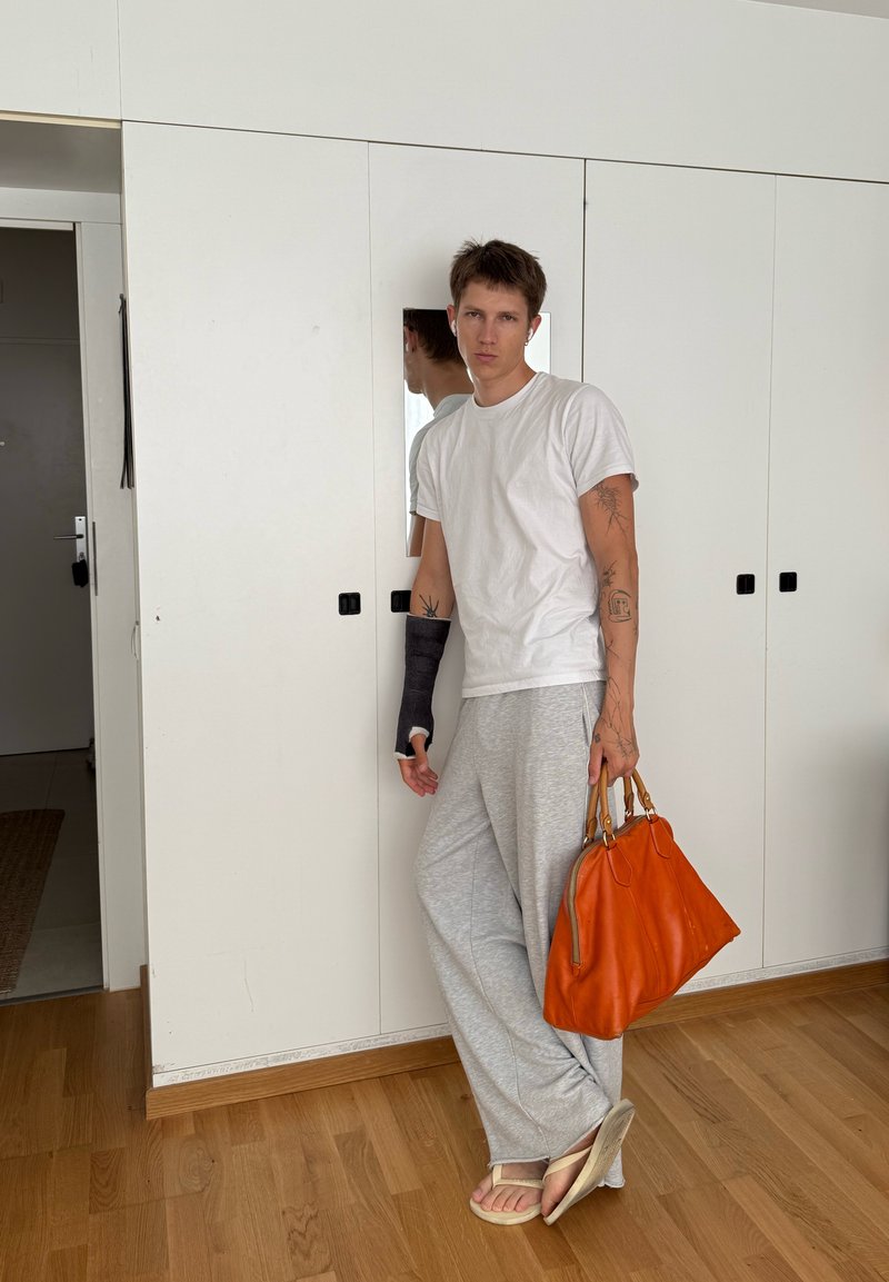 Jeune homme avec des tatouages et une attelle noire au poignet, appuyé contre un placard blanc, tenant un sac à main orange et portant un pantalon gris et des sandales.