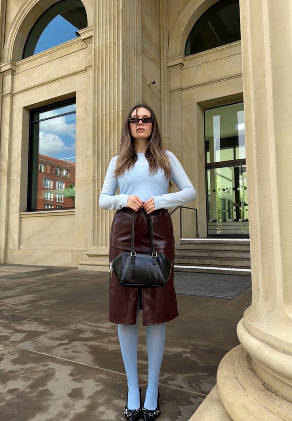 Femme debout devant un bâtiment en pierre beige avec des colonnes, portant des lunettes de soleil, un haut bleu clair, une jupe marron foncé, des collants bleu clair, et tenant un sac à main noir.