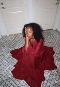 Femme aux cheveux bouclés assise sur un sol carrelé à motifs, portant une longue robe rouge fluide, reposant sa tête sur ses mains entre deux portes blanches fermées.