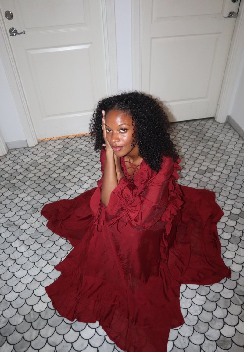 Vrouw met krullend haar zit op een vloer met patroontegels, draagt een vloeiende lange rode jurk en rust haar hoofd op haar handen tussen twee witte gesloten deuren.