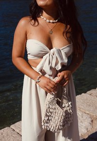 Beige, trägerloses Top mit Bindeband, kombiniert mit einem fließenden Rock. Hält eine geflochtene Tasche mit weißen Muscheln und trägt eine Muschelkette.