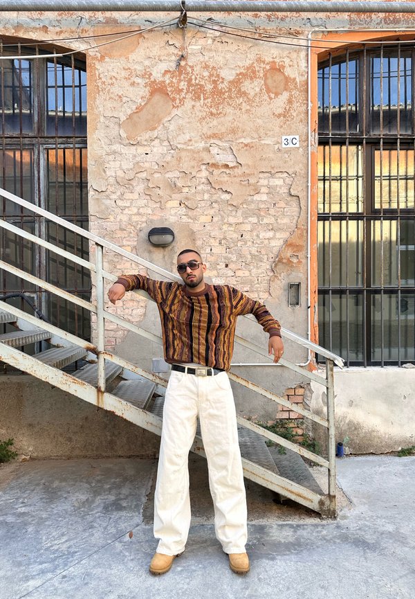 Maglione a righe verticali marroni e gialle, pantaloni bianchi a gamba larga e stivali beige. Scale in metallo e un muro di mattoni texturizzato sullo sfondo.