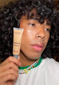 MÁDARA Skinonyms ημι-ματ μακιγιάζ με πεπτίδια σε μπεζ σωλήνα, το κρατάει ένα άτομο με σγουρά μαλλιά, φορώντας πολύχρωμο κολιέ με χάντρες.