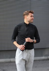 Top atletico nero con zip, pantaloni della tuta grigi, mentre tiene in mano una bottiglia d'acqua nera. Lo sfondo presenta listelli metallici grigi orizzontali.