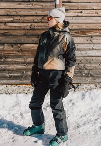 Tenue de ski comprenant une veste noire et beige, un pantalon noir et des chaussures de ski vertes. Le matériau semble isolé avec des poches et des gants. Fond enneigé.