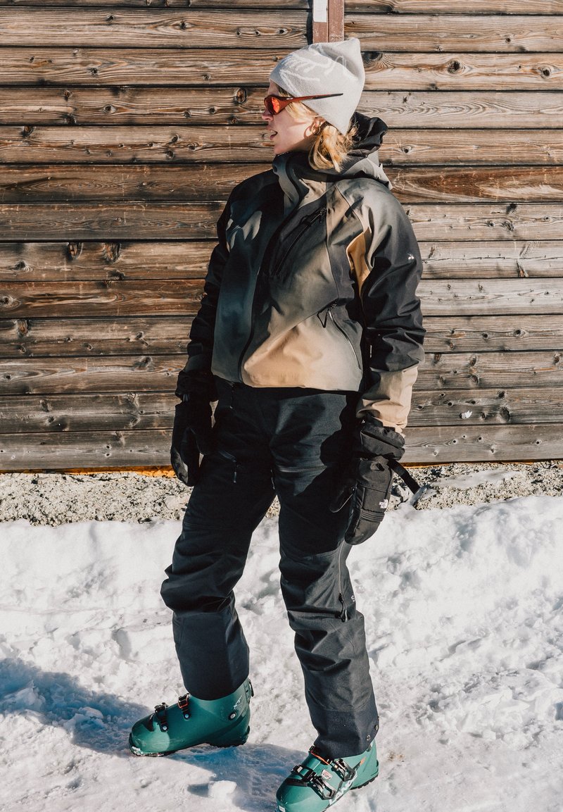 Tenue de ski comprenant une veste noire et beige, un pantalon noir et des chaussures de ski vertes. Le matériau semble isolé avec des poches et des gants. Fond enneigé.