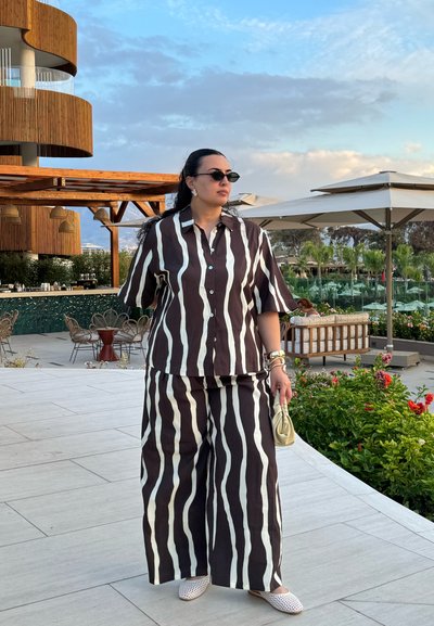 Camisa de botones de manga corta a rayas marrón y blanca con pantalones anchos a juego. Los accesorios incluyen gafas de sol y un pequeño bolso.