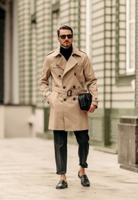 Beige trenchcoat med dubbelknäppta knappar, bälte och axellappar. Kombinerad med svart polotröja, mörka byxor och loafers.