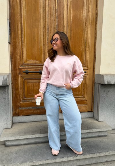 Joven mujer con sudadera rosa y vaqueros anchos azul claro, sosteniendo una taza de café, de pie en unos escalones frente a una gran puerta de madera.
