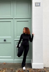 Schwarzes Sportoutfit mit langen Ärmeln und enganliegenden Hosen. Heller grüner Türhintergrund, mit einem dunklen runden Türknauf und der Hausnummer 28.