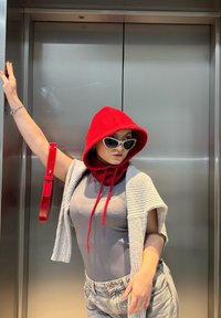 Rød hættetrøje med snore, tætsiddende grå top og lyseblå jeans. Katteøje-solbriller. Holder et rødt bælte mod en metallisk elevator.