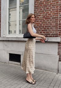 Beige prikkete undertøy med en flytende design, kombinert med en hvit topp og svart håndveske. Flate svarte sandaler med blomsterdetaljer fullfører stilen.