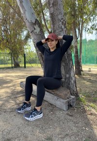 Noir haut à manches longues, leggings noirs et chaussures de sport grises avec des semelles blanches. Le chapeau est rouge avec un logo. Debout à côté d'un arbre dans un parc.