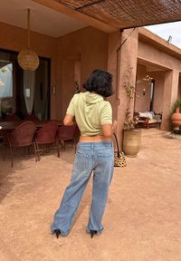 Grüner, kurz geschnittener Kapuzenpullover, hellblaue Jeans mit weiter Beinweite und schwarze Stiefel mit Absatz. Dazu eine gemusterte Handtasche. Außenbereich mit Terrakottatönen.