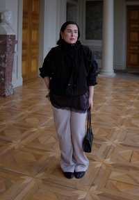 Veste noire surdimensionnée à col haut, portée sur un haut orné de dentelle. Pantalon beige clair et chaussures plates noires complètent la tenue.