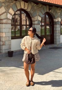 Cárdigan de punto texturizado en color crema con botones redondos, falda corta de cuadros y bolso marrón. Zapatos planos, ambiente al aire libre con arquitectura de piedra.