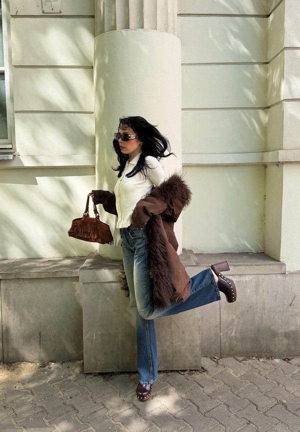 Femeie cu păr închis la culoare purtând ochelari de soare, top alb, palton de blană maro, blugi albaștri și saboți ridică un picior în timp ce ține o geantă maro cu franjuri.