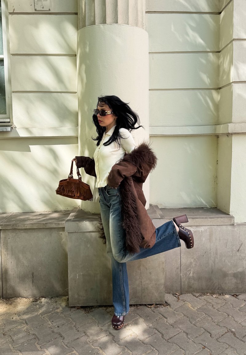 Γυναίκα με σκούρα μαλλιά που φοράει γυαλιά ηλίου, λευκό τοπ, καφέ παλτό με γούνα, μπλε τζιν και ξύλινα παπούτσια σηκώνει ένα πόδι ενώ κρατά καφέ τσάντα με κρόσσια.