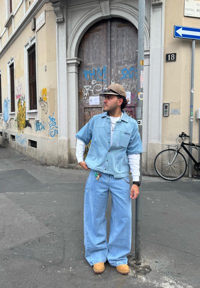 Camisa de mezclilla azul claro con mangas cortas, superpuesta sobre una camiseta blanca de mangas largas. Jeans azul claro de corte holgado y zuecos en color tostado. Gorra con estampado.