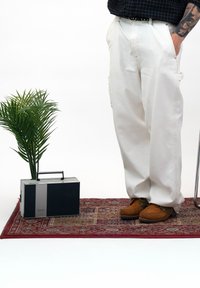 Personne portant un pantalon blanc et des chaussures marron se tenant sur un tapis à motifs à côté d'une radio Philips vintage et d'une plante verte en pot.