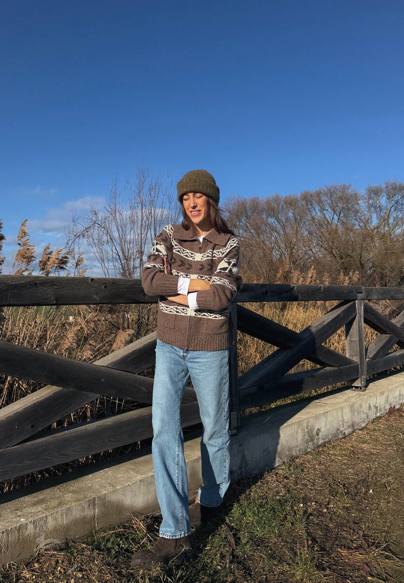 Femme portant un pull marron à motifs, un jean bleu et un bonnet, debout les bras croisés près d'une clôture en bois dans un espace herbeux en plein air.