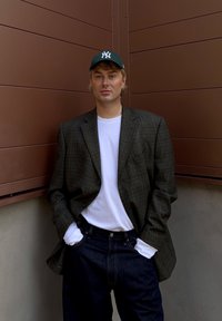 Blazer de cuadros en verde oscuro y marrón, camiseta blanca, jeans oscuros y una gorra de béisbol verde oscuro con letras blancas. Fondo neutro.