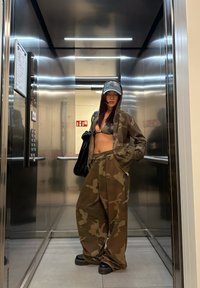 Kreklu ar kamuflāžas rakstu un biksēm brūnos toņos, ar īsu topu un cepuri, apvienots ar melnām platformas kurpēm spoguļliftā.