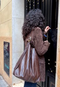 Femme aux cheveux bouclés portant une veste en daim marron et un grand sac en cuir déverrouillant une porte en métal noir dans une rue de la ville.