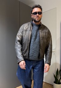 Homme portant des lunettes de soleil noires, un pull gris, une veste en cuir marron et un jean bleu, debout à l'intérieur près d'une armoire grise et d'une porte blanche.