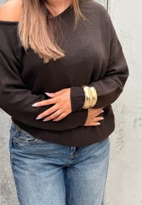 Vrouw in een donker off-shoulder trui, blauwe jeans, gouden armbanden en een zilveren ring met een lichte manicure, staande tegen een effen muur.