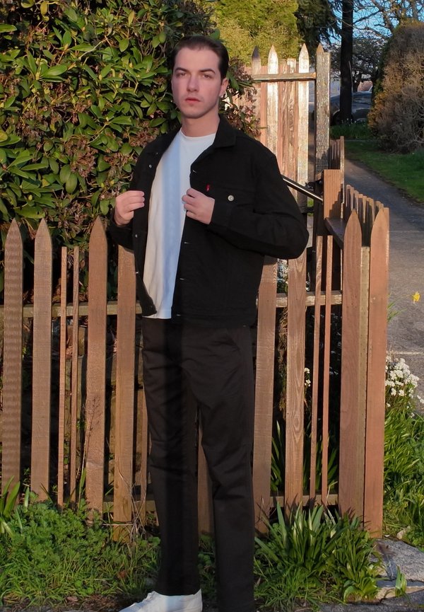 Ung mann i svart jakke og bukser justerer jakken mens han står ved et gjerde av tre med grønt i sollys.