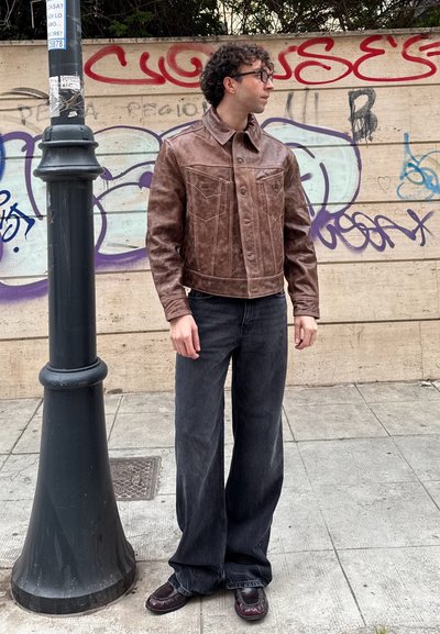 Chaqueta de cuero marrón corta con un diseño de bolsillo frontal, combinada con jeans oscuros de pierna ancha y zapatos marrones, de pie al lado de un farol.
