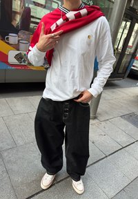 Personne portant une chemise blanche à manches longues, un jean noir ample, des baskets blanches et un vêtement rouge drapé sur les épaules, debout sur un trottoir urbain près d'un arrêt de bus.