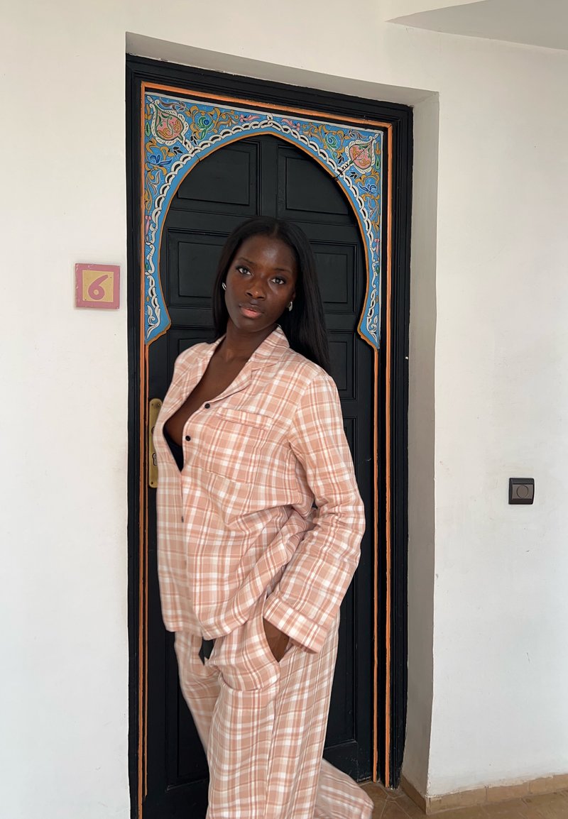 Femme en pyjama à carreaux marron clair pose les mains dans les poches devant une porte noire avec un arc décoratif coloré.