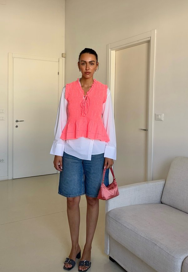 Kvinde stående indendørs iført en koralærmet top over en hvid bluse, denimshorts, sorte sandaler, og holder en lille lyserød taske.