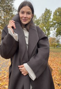 Tmavě hnědý oversize vlněný kabát s velkým límcem, žebrovanými krémovými manžetami a bočními kapsami, vrstvený p�řes světlý svetr. V pozadí podzimní listí.