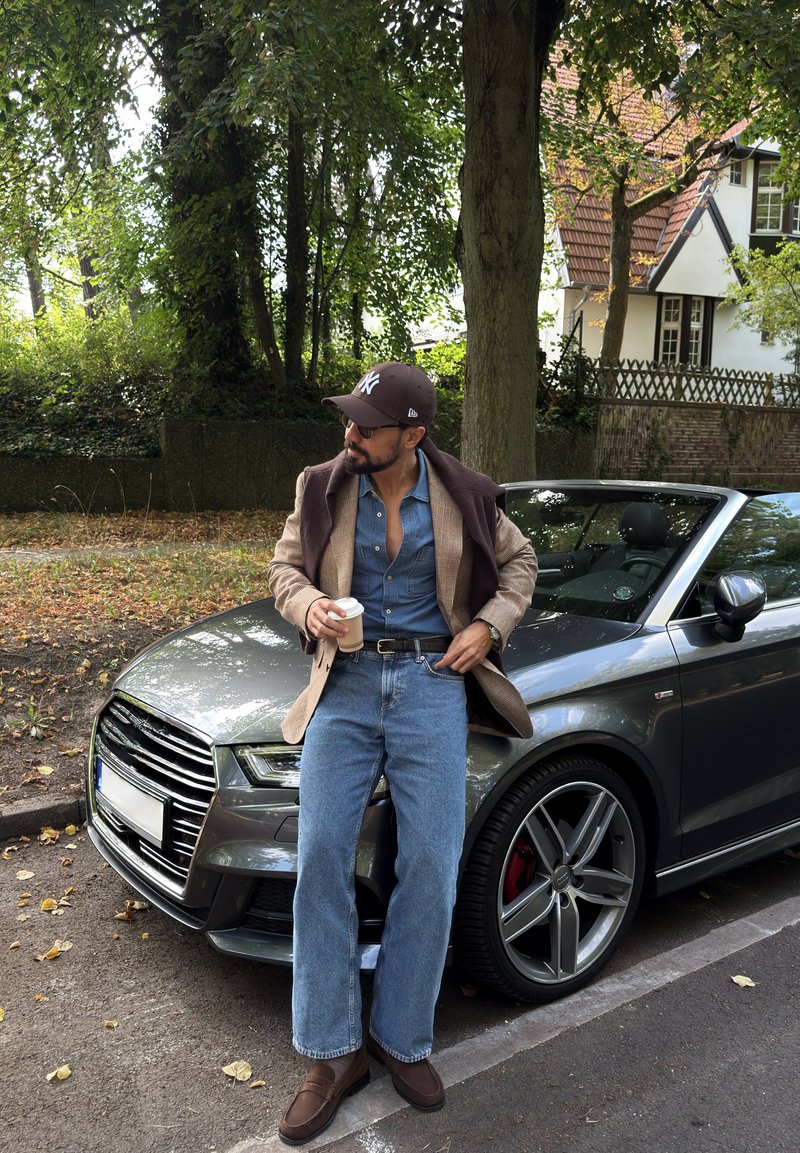 Un hombre con barba, gorra y gafas de sol sostiene un café para llevar junto a un coche gris aparcado en una calle residencial arbolada.