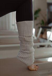 Paljas jalka, jossa tummat kynsilakat, pukeutuneena löysiin kermaisiin neulottuihin säärystimiin, varpaillaan pehmeästi valaistussa sisätilassa pehmustetulla pinnalla.