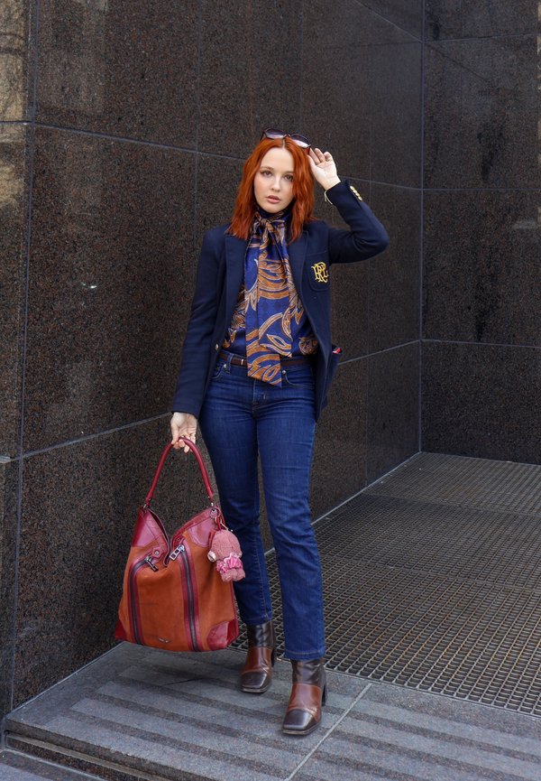 Kvinne med rødt hår iført mønstret skjerf, marineblå blazer, jeans og brune støvler, holder en stor brun og rød håndveske nær en mørk steinvegg.