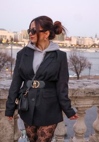 Blazer preto oversized sobre um hoodie cinza, meias-calças com padrão renda, fivela dourada e óculos de sol. Uma mini bolsa preta pendurada em um ombro.