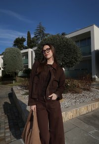 Jeune femme portant des lunettes et une tenue marron, tenant un grand sac beige, debout sur un chemin près de bâtiments blancs modernes et d'arbres taillés en pleine journée.