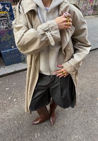 Beežist trentšmantel halli lukustatava kapuutsiga dressipluusi kohal, kombineeritud tumedate teksapükstega ja teravate pruunide saabastega. Sõrmi kaunistavad mitmed sõrmused.